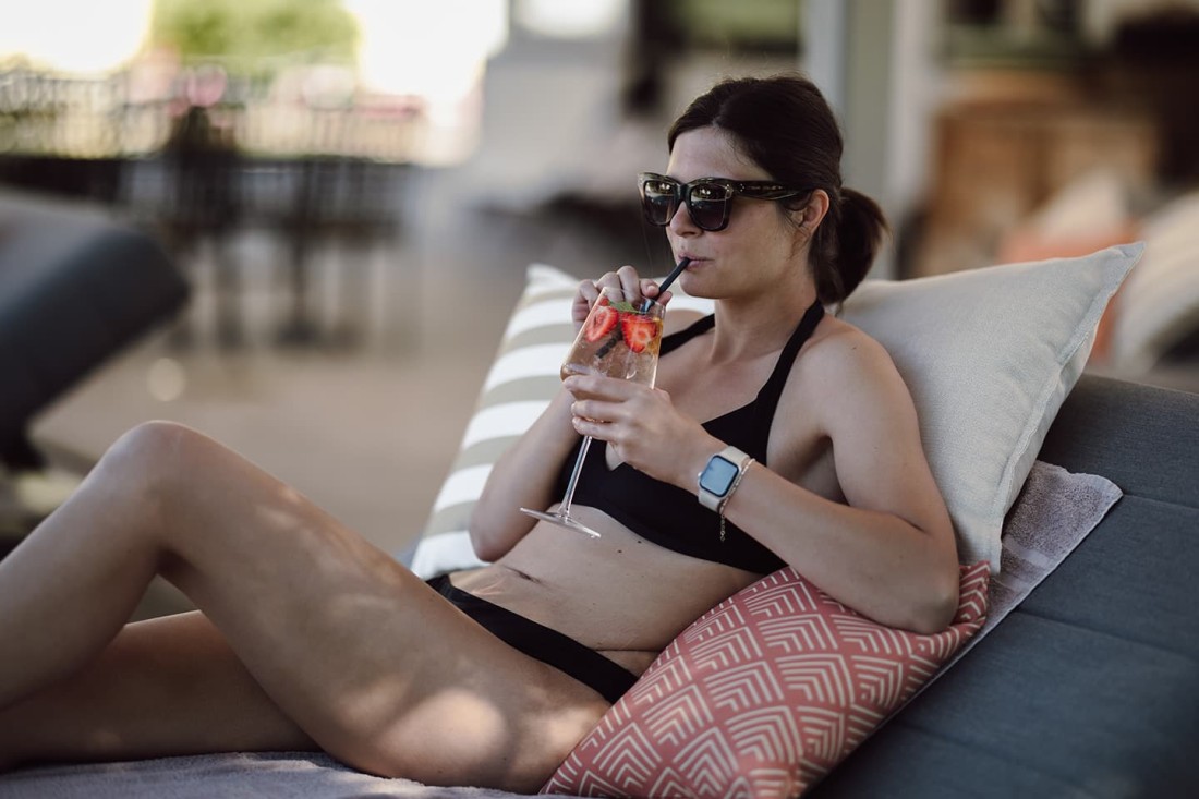 Frau in Bikini liegt auf Liegestuhl und trinkt durch einen Strohhalm aus einem Cocktailglas
