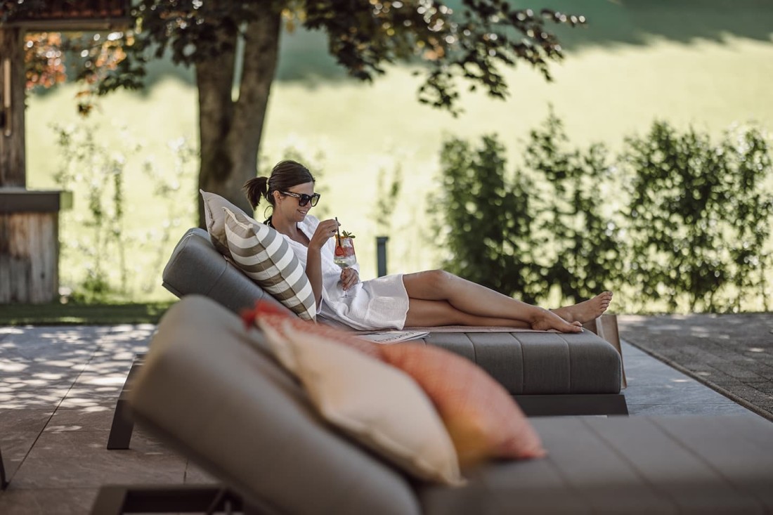 Eine Frau entspannt auf einer Outdoor-Couch und h&auml;lt einen Drink in der Hand beim Relaxan am Pool im Hotel loj
