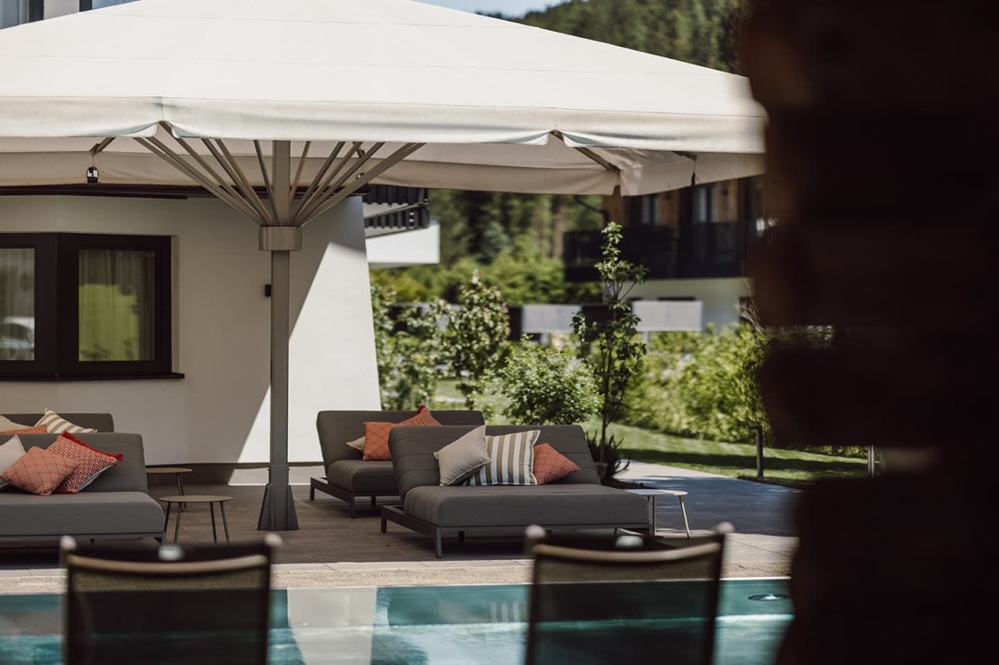 Zwei graue Liegen mit Kissen unter einem gro&szlig;en Sonnenschirm neben dem Hotelpool vom loj
