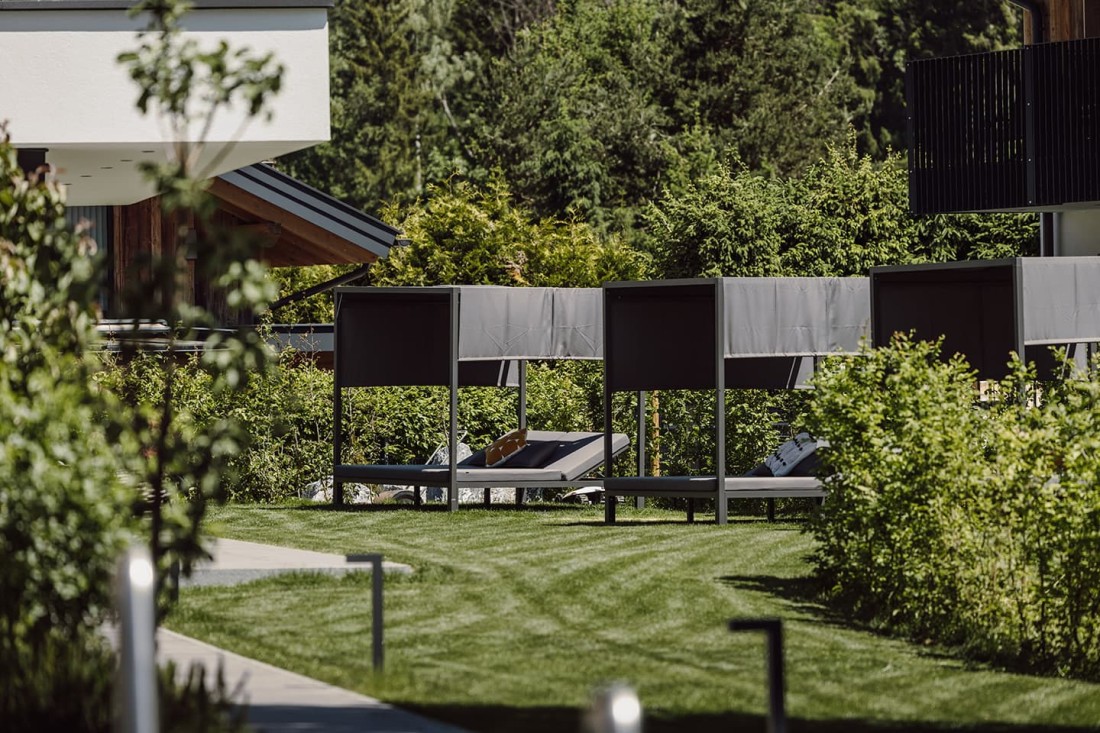 Sonnenliegen mit &Uuml;berdachungen auf einer Wiese vor gr&uuml;ner Hecke und B&auml;umen im Hotelgarten vom loj
