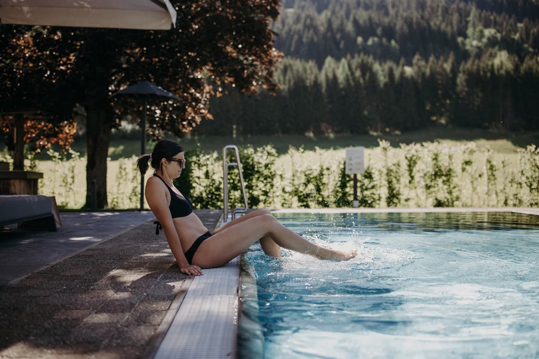 Eine Frau im Bikini sitzt am Pool und planscht mit den F&uuml;&szlig;en im Wasser